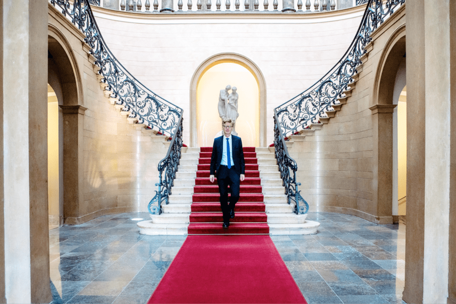 Alt Tag: Ein Mann in Anzug und blauer Krawatte läuft auf einem roten Teppich eine Treppe herunter
